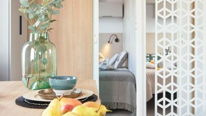 Una habitación con una mesa de madera, un florero con follaje, un tazón y una cama con una manta gris.