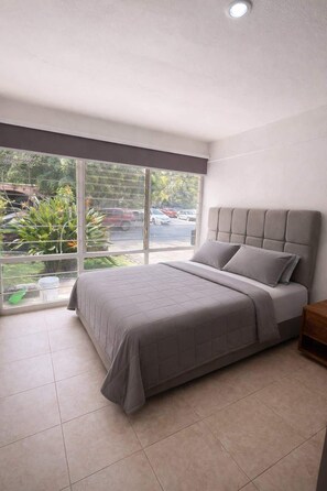 Ein Schlafzimmer mit großem Fenster, einem Bett mit grauer Bettwäsche und einem Nachttisch.
