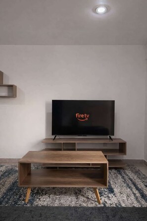 Ein modernes Wohnzimmer mit einem Flachbildfernseher an einer Holzkonsole, einem dunkelgrauen Sofa und einem gemusterten Teppich.
