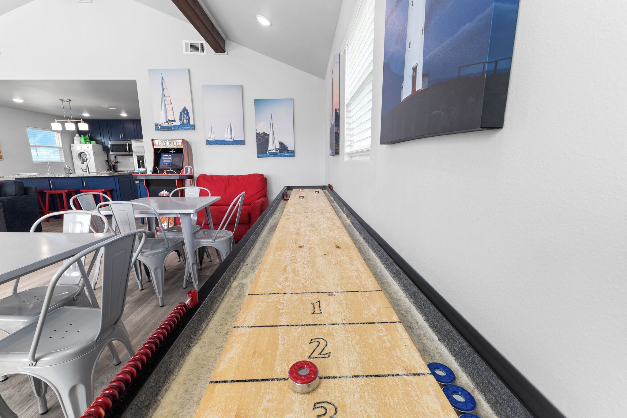 A shuffleboard table in a room with a kitchen area, dining chairs, and artwork on the walls.