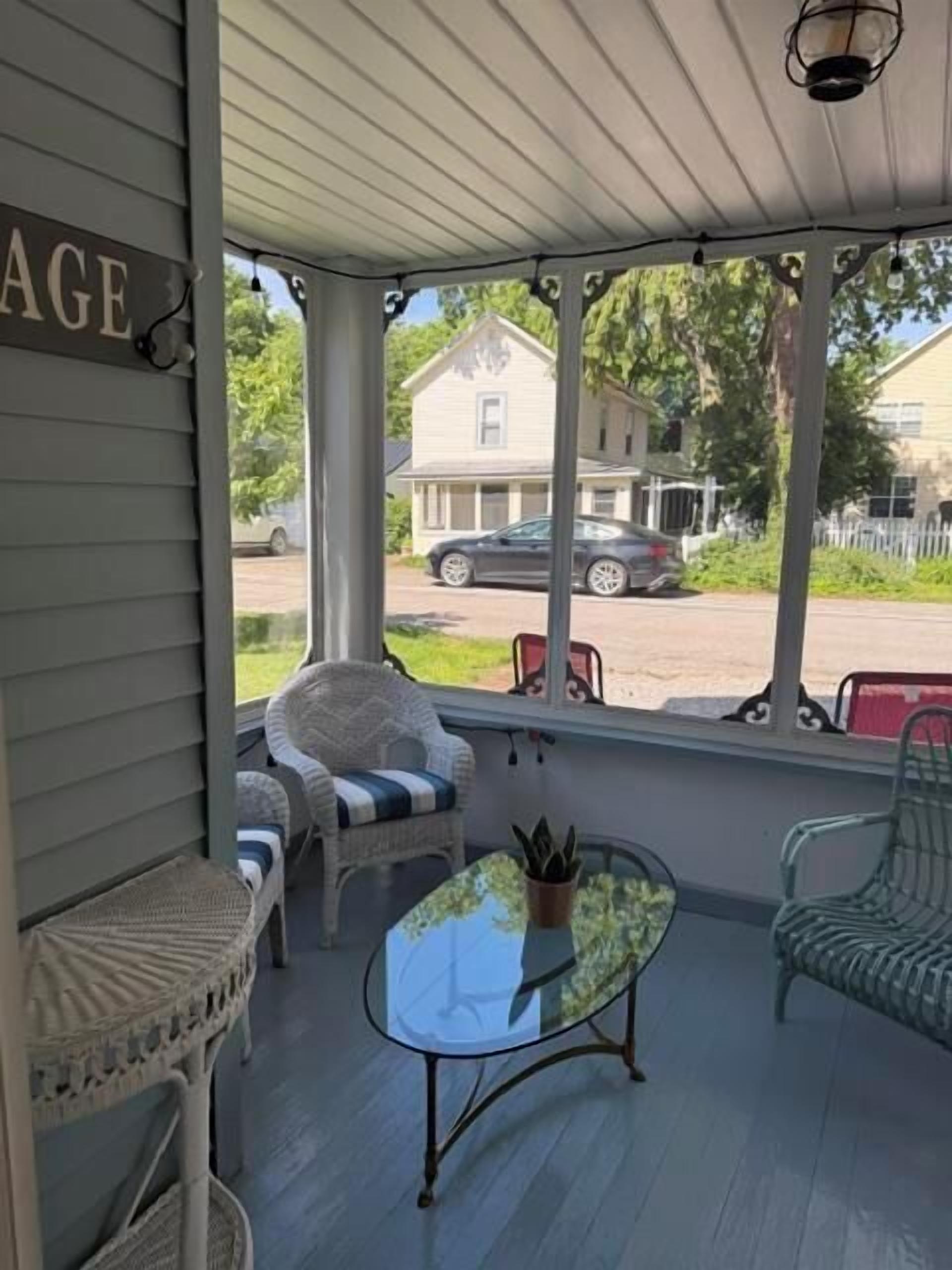 A covered porch with wicker furniture, a glass table, and a view of a house and street.