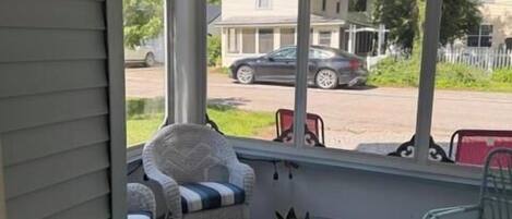 A covered porch with wicker furniture, a glass table, and a view of a house and street.