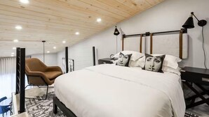 Una habitación de hotel moderna con techo inclinado, una cama grande con ropa blanca, un sillón marrón, un escritorio y una lámpara.