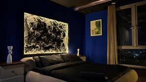 A modern bedroom with a large bed, a wall-mounted light feature, and a window with a night view. - luxury 1 bed apartment with indoor cinema,starlights & cityview in centre of DHA (Lahore)