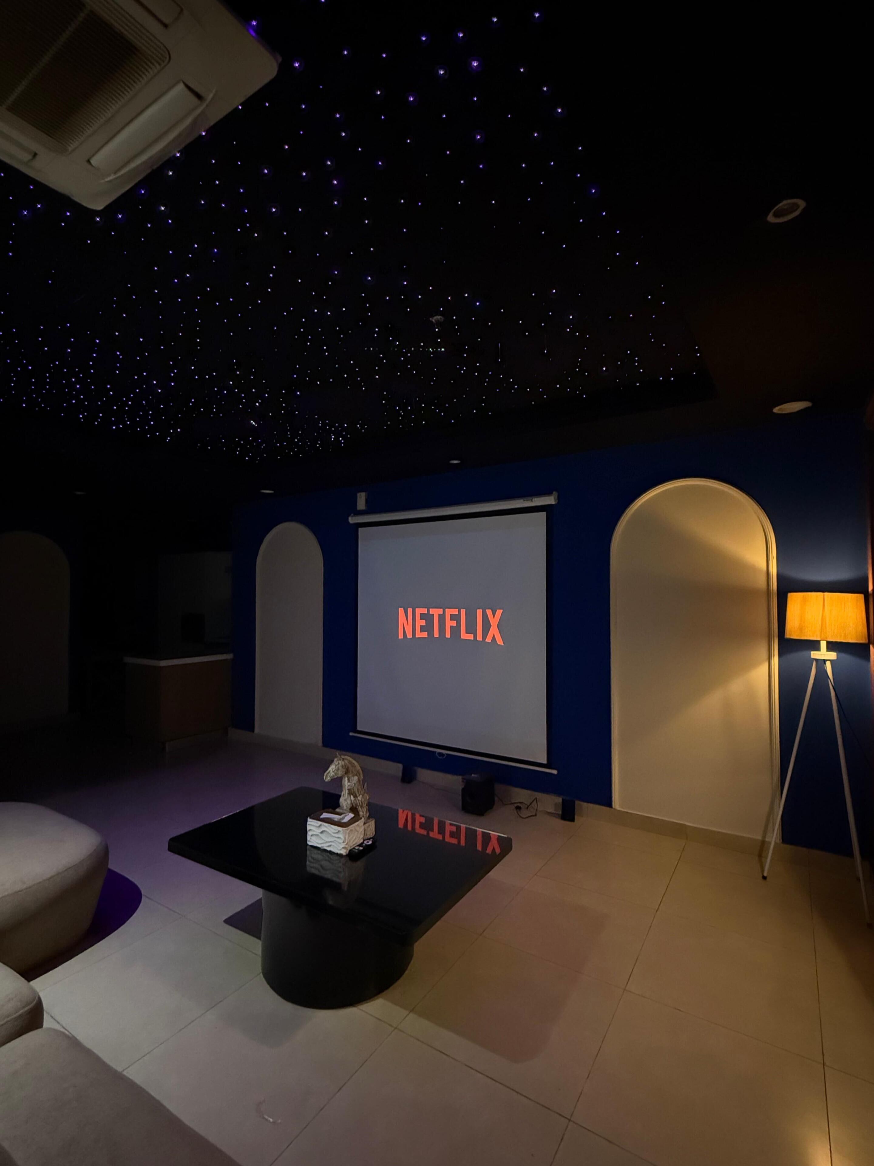 A room with a starry ceiling, a large screen displaying 'NETFLIX,' a small table with a sculpture, and a lamp.