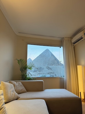 A room with a view of a pyramid, a sofa, a plant, and an air conditioning unit. - Pyramids View Suite (محافظة الجيزة)