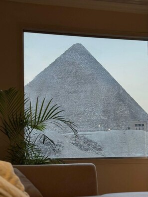 A view of a pyramid through a window, with a plant to the left and a chair in the foreground. - Pyramids View Suite (محافظة الجيزة)