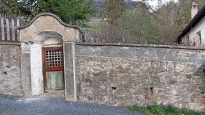 Un mur de pierre surmonté d’un portail en métal, une clôture en bois et un bâtiment à l’architecture traditionnelle.