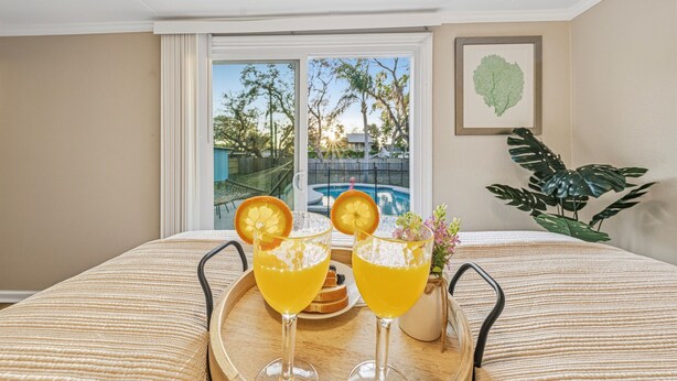 A room with a bed, a tray with two drinks, a plant, and a window with a view of a pool and trees. - Cozy 3BR Clearwater Home w/ Private Pool & Backyard Oasis (Clearwater)