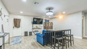 Uma sala de estar moderna com um sofá azul, uma mesa de jantar com quatro cadeiras, uma TV de tela plana na parede e um ventilador de teto.