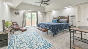 Quarto com cama, escrivaninha, cadeira, banco, ventoinha no teto e uma janela grande.