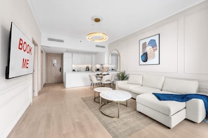A modern living room with a sofa, coffee table, and a kitchen in the background. - Free Housekeeping Visits During Your Stay - Stayshort - Elegant 1 Bedroom in JVC Sleeps 4 With Balcony (Dubai)