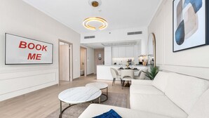 A modern living room with a sofa, a coffee table, a TV displaying 'BOOK ME', and a kitchen in the background. - Free Housekeeping Visits During Your Stay - Stayshort - Elegant 1 Bedroom in JVC Sleeps 4 With Balcony (Dubai)