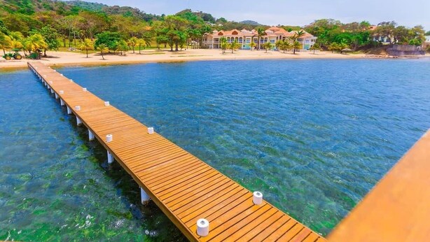Eine Holzstege, der sich in klares, blaues Wasser erstreckt, mit einem Strand und Häusern im Hintergrund.