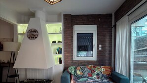 Ein Wohnzimmer mit Couchtisch aus Holz, einem blauen Sofa mit bunter Decke, einem weißen Kamin und einem großen Fenster mit Vorhängen.