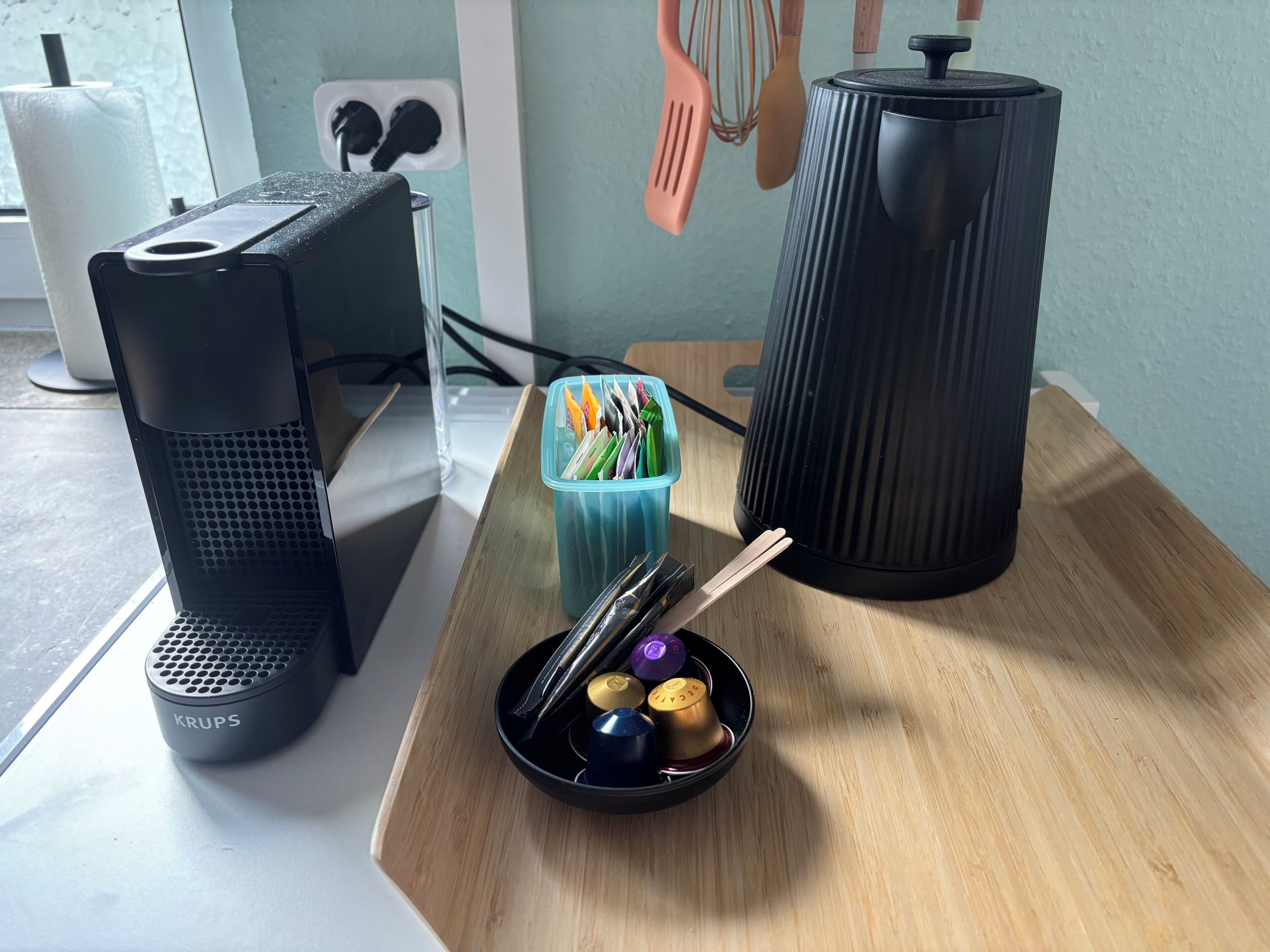 Ein Computerarbeitsplatz mit einer Krups-Kaffeemaschine, einer Aufbewahrungsbox für Besteck und einem zylindrischen Gerät auf einem Holztisch.