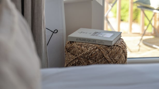 Una habitación con cama, ventana con vista a un balcón y un libro titulado 'A Frame for Life' sobre un taburete tejido.