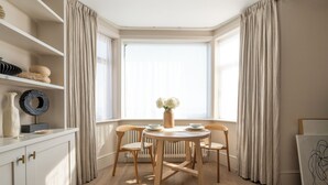 Un comedor con una mesa redonda, cuatro sillas, un jarrón con flores, un armario blanco con estantes y una ventana con cortinas.