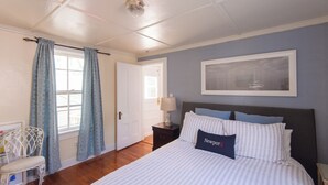 Una camera da letto con un letto, un comodino, una sedia, una finestra con tende e un quadro appeso al muro.