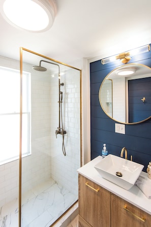 Une salle de bain avec une douche à parois de verre, un lavabo blanc, un meuble-lavabo en bois et un miroir rond.