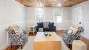 Una stanza con soffitto in legno, due poltrone a motivi decorativi, un divano scuro, un tavolino in legno e una porta bianca.