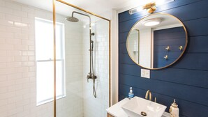 Un bagno con doccia vetrata, lavabo bianco con accessori dorati e specchio tondo su una parete blu scuro.