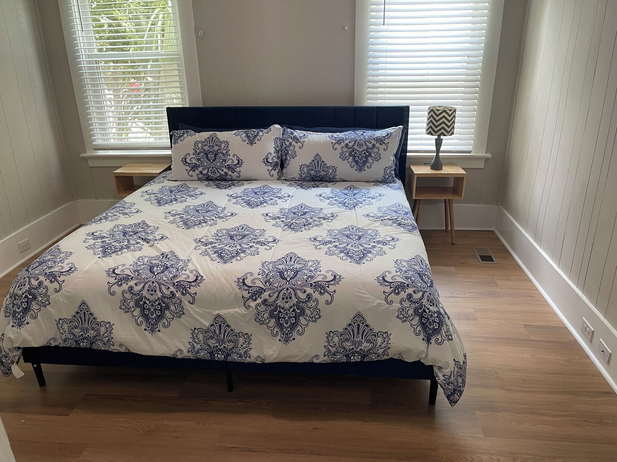 A bed with a patterned bedspread, two bedside tables, and a window with blinds.