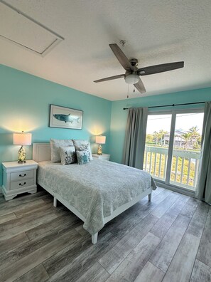 A bedroom with a bed, two bedside tables, a ceiling fan, a window with curtains, and a view of the outdoors.