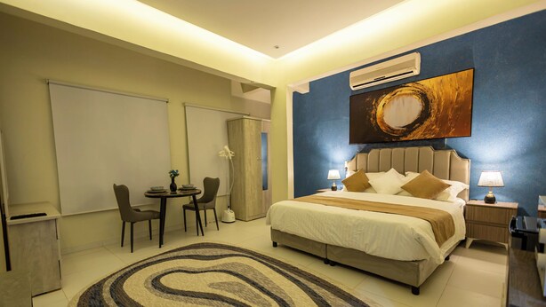 A hotel room with a bed, bedside tables, a desk with chairs, a large rug, and a painting on the wall. - Self Checkin Studio - Olaya 203 (Riyadh)
