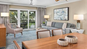 Habitación de hotel con zona de comedor, un conjunto de sofás, ventilador de techo y un gran ventanal con cortinas.