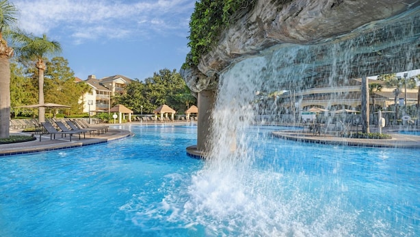Una piscina con cascada, sillas de descanso y un edificio al fondo.