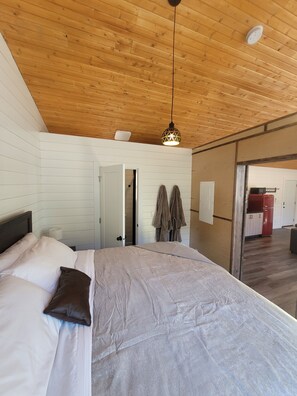 Camera da letto con soffitto in legno, un letto con biancheria da letto bianco e marrone, una lampada a sospensione e una porta scorrevole che dà su un'altra stanza.