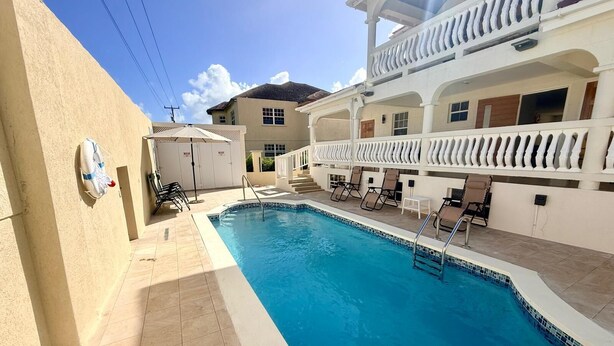 A swimming pool with lounge chairs, a covered area, and a building with a balcony. - 1BR NEWLY REFURBISHED Apartment | Pool | Minutes to Oistins (Oistins)