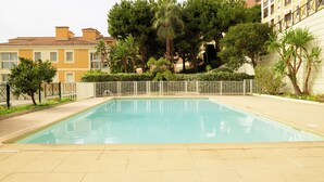 Una piscina rettangolare con bordo piastrellato, circondata da una recinzione bianca, alberi e edifici residenziali.