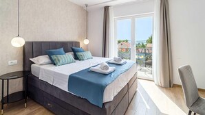 A modern hotel room with a large bed, a small side table, a chair, and a balcony with a view.