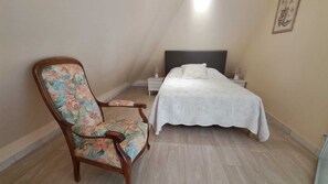 A bedroom with a bed, a nightstand, a chair with floral upholstery, and a wall-mounted artwork. - Charming apartment near Deauville (Blonville-sur-Mer)