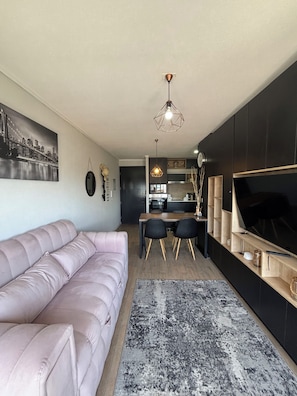 Un salon moderne avec un canapé rose, un coin repas avec des chaises, une cuisine en arrière-plan et une grande télévision sur le côté droit.