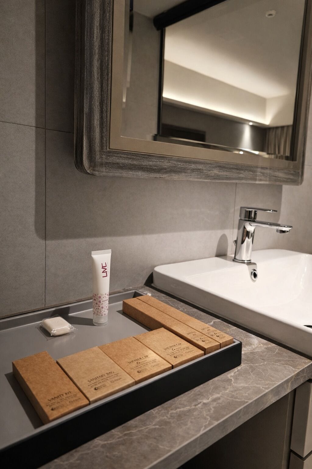 A bathroom with a large mirror, a white sink, and a countertop with a tube of lotion and boxes.