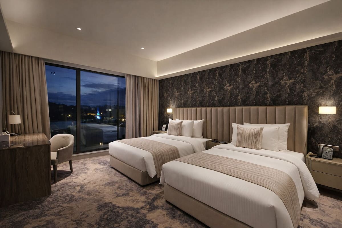 Ein Hotelzimmer mit zwei Betten, einem Schreibtisch, einem Sessel und einem großen Fenster mit Aussicht. Die Wand ist dunkel gemustert.