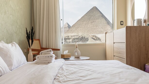 Een hotelkamer met een bed, een raam met uitzicht op een piramide, een houten dressoir en een kleine tafel met een vaas en een afstandsbediening.
