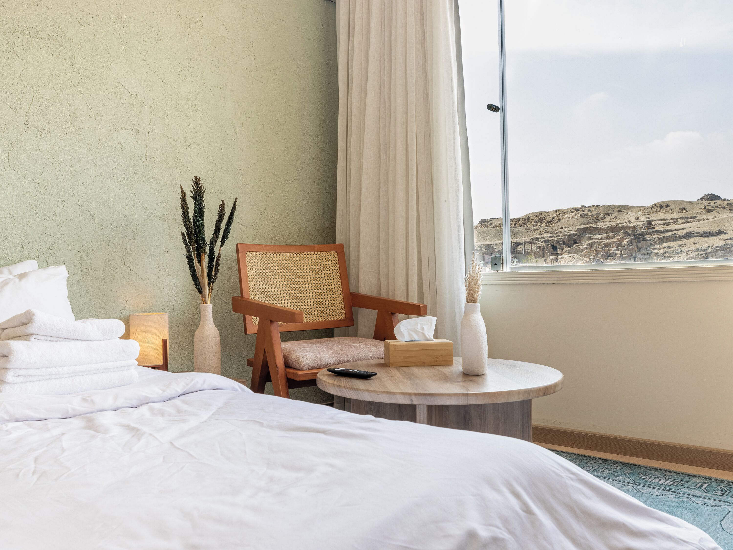 A hotel room with a bed, a wooden chair, a small round table, a vase with dried plants, and a view of a desert landscape.