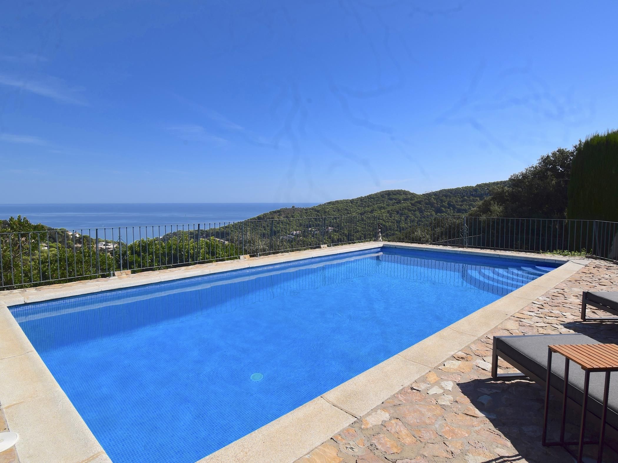 A rectangular swimming pool with a clear blue interior, surrounded by a stone deck and metal railing, overlooking a coastal landscape.