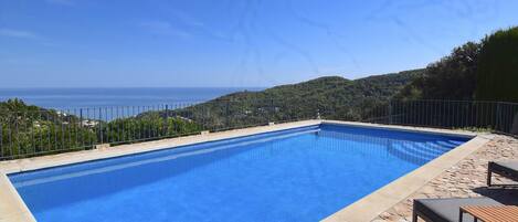 A rectangular swimming pool with a clear blue interior, surrounded by a stone deck and metal railing, overlooking a coastal landscape.