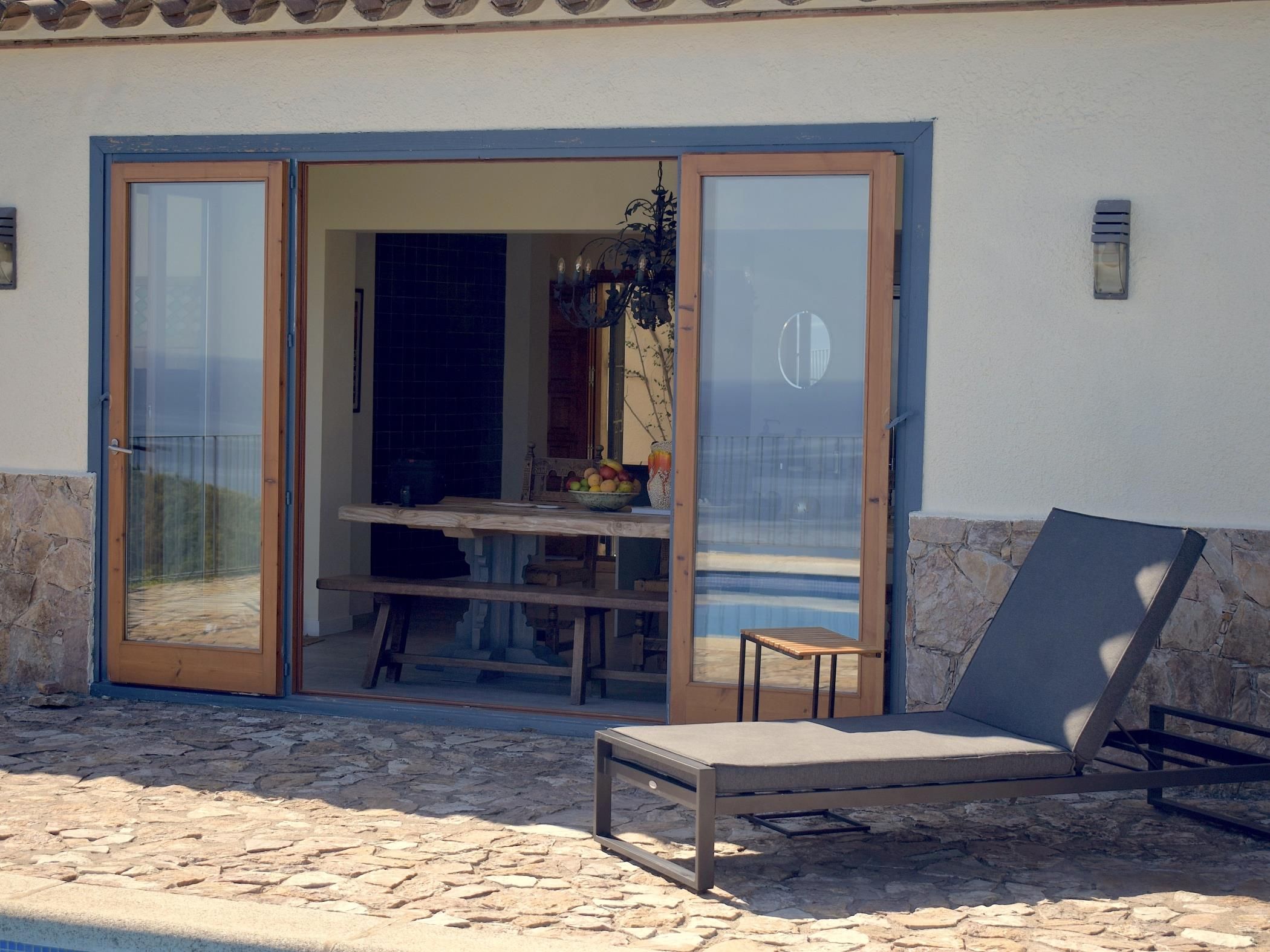 A patio with a stone floor, a wooden table, a chair, and a glass door leading to an interior space with a tiled wall and a hanging plant.