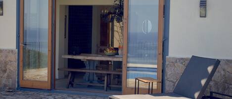 A patio with a stone floor, a wooden table, a chair, and a glass door leading to an interior space with a tiled wall and a hanging plant.