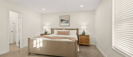 A bedroom with a bed, bedside tables, a painting, and a window with blinds.