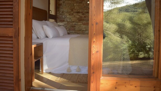 Une chambre à coucher avec un lit, une tête de lit en bois, un mur en pierre et une vue sur les arbres par la fenêtre.