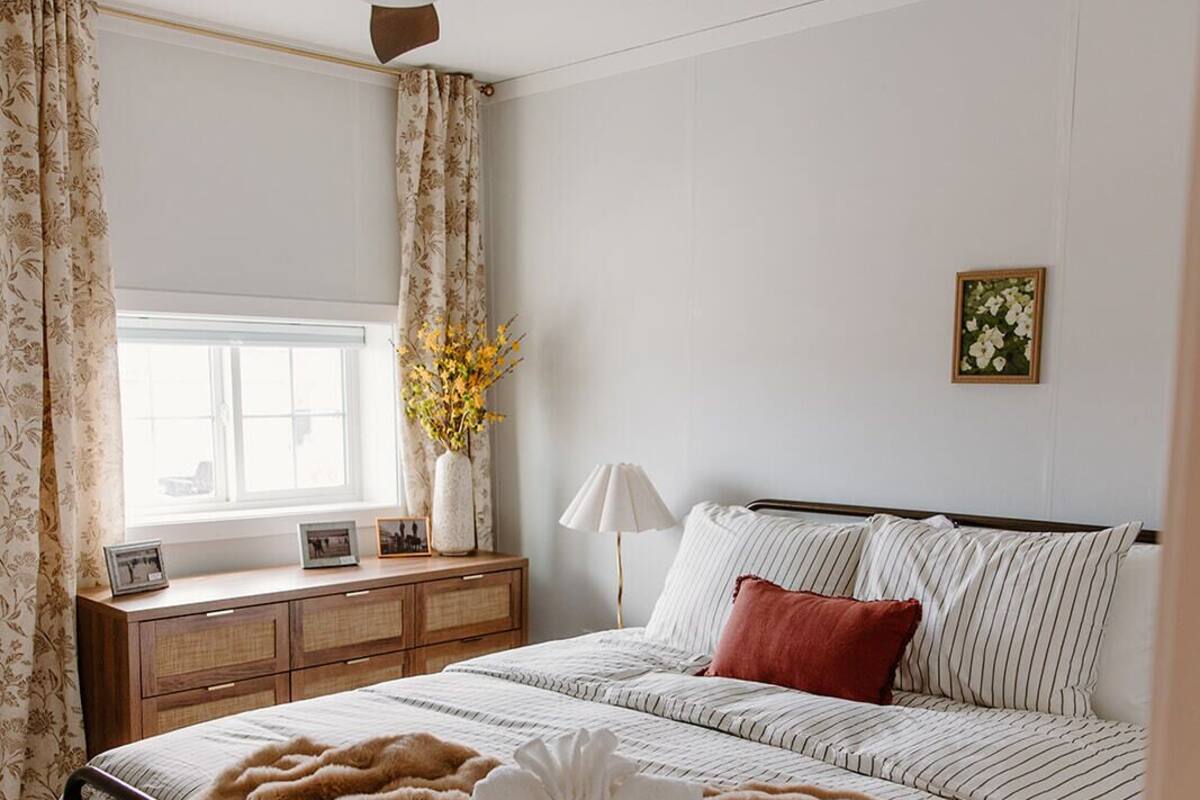 Un dormitorio con cama, ventilador de techo, mesita de noche, lámpara, ventana con cortinas y un cuadro en la pared.