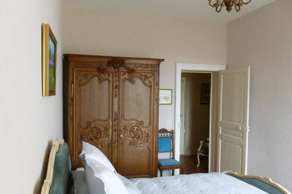 A bedroom with a bed, a wooden wardrobe, a chair, and a door leading to another room.