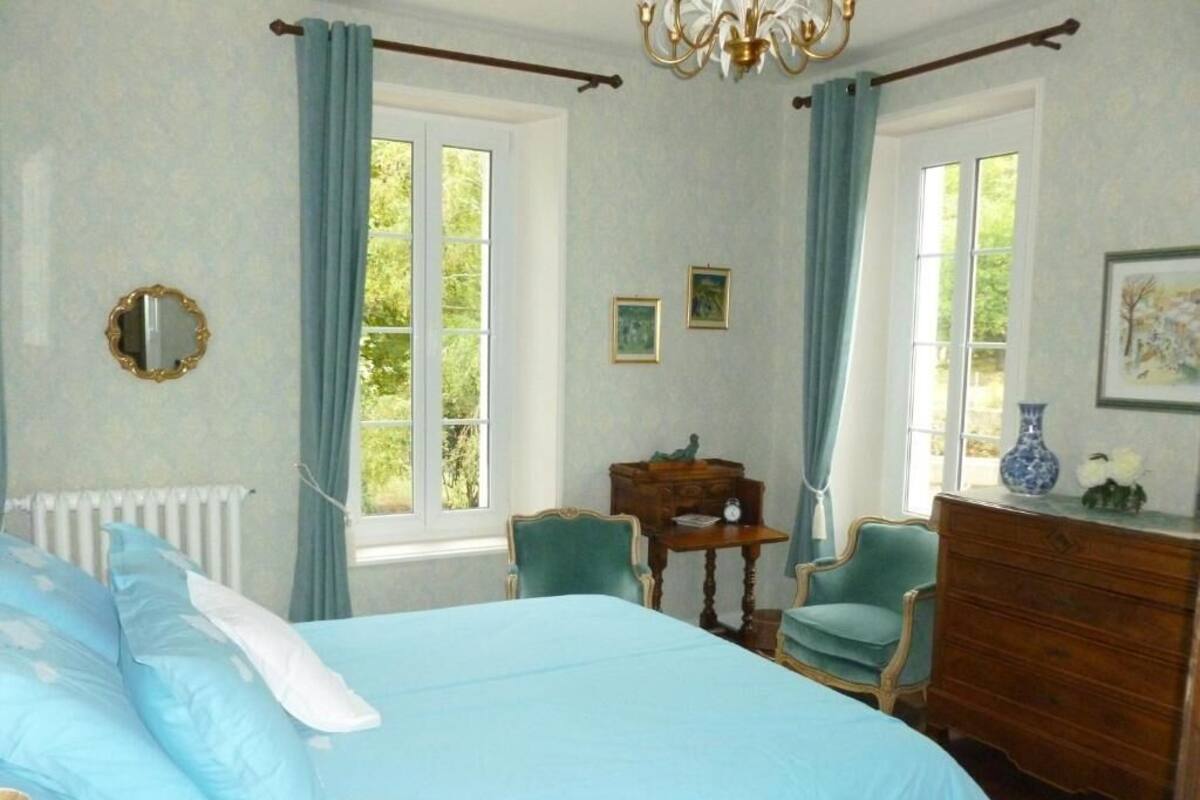 A bedroom with a bed, two chairs, a dresser, a mirror, and a window with curtains.